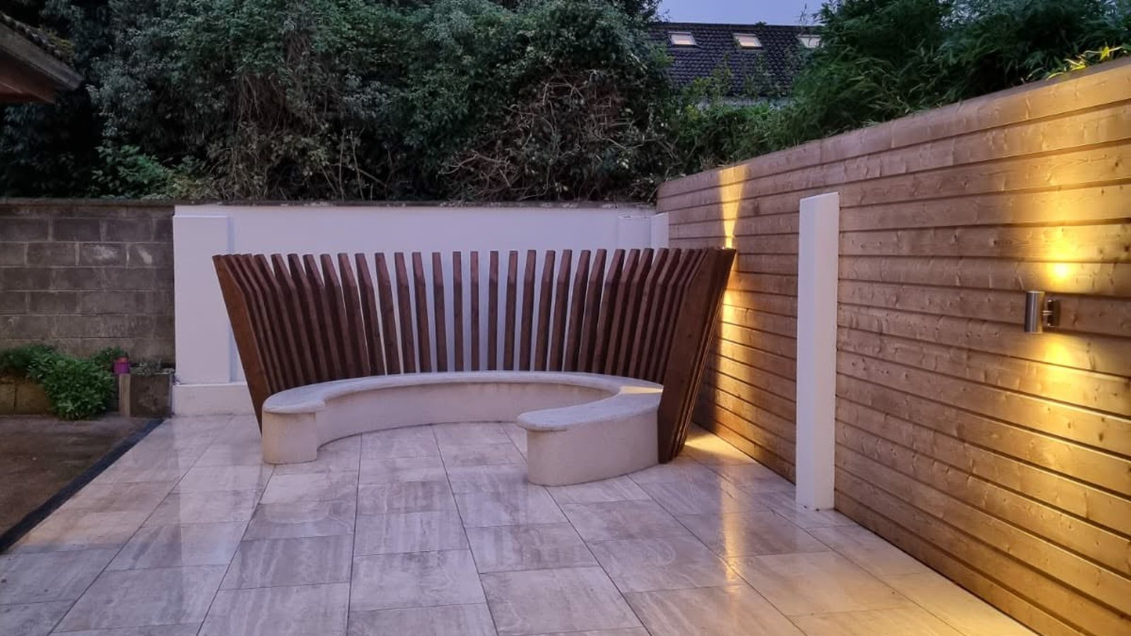 A finished patio at dusk: a curved seat with a slatted timber back, uplit timber wall and porcelain paving